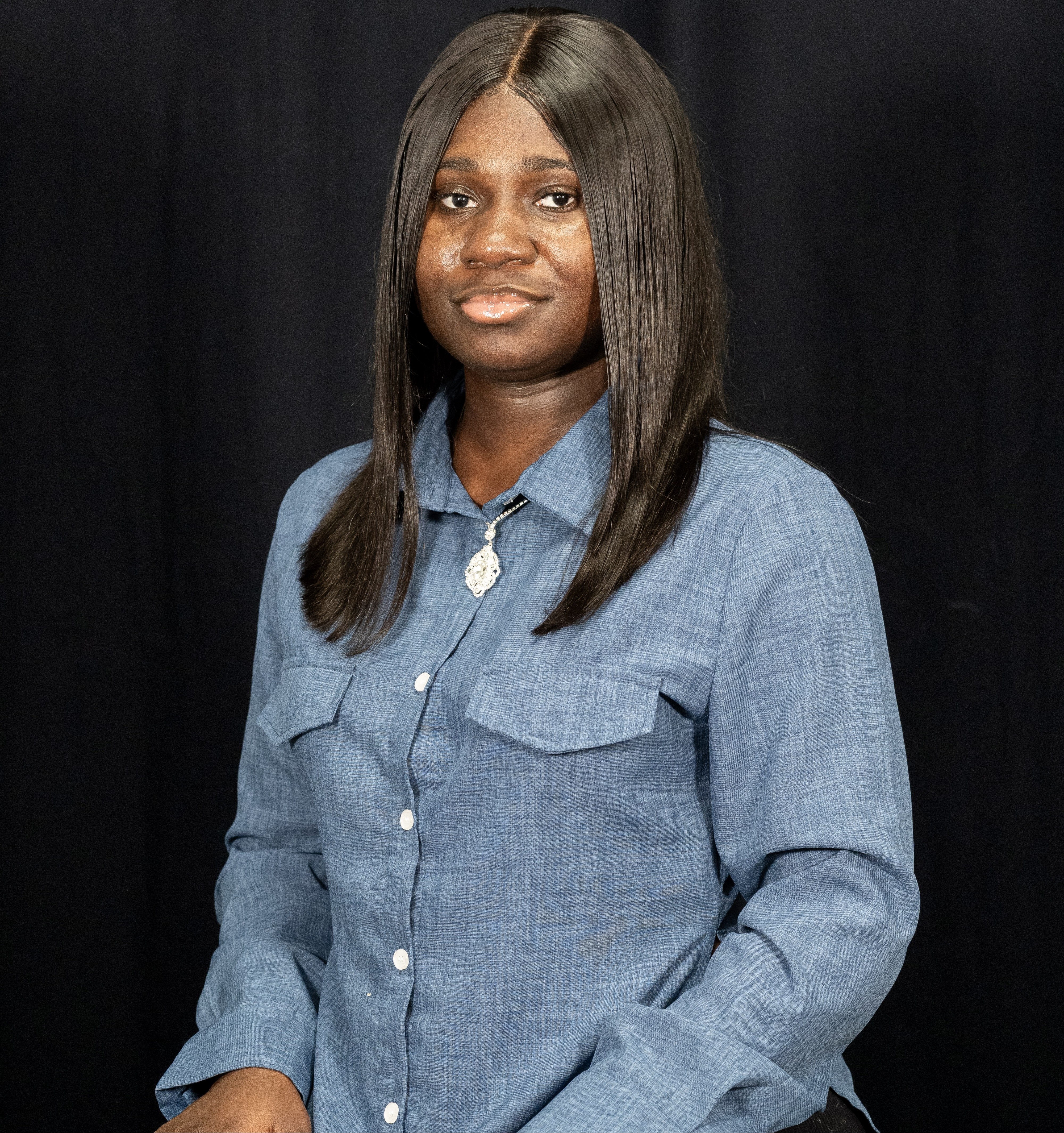 Photograph of Nana Gyamfi facing forward wearing a blue shirt and silver pendant in front of a black background