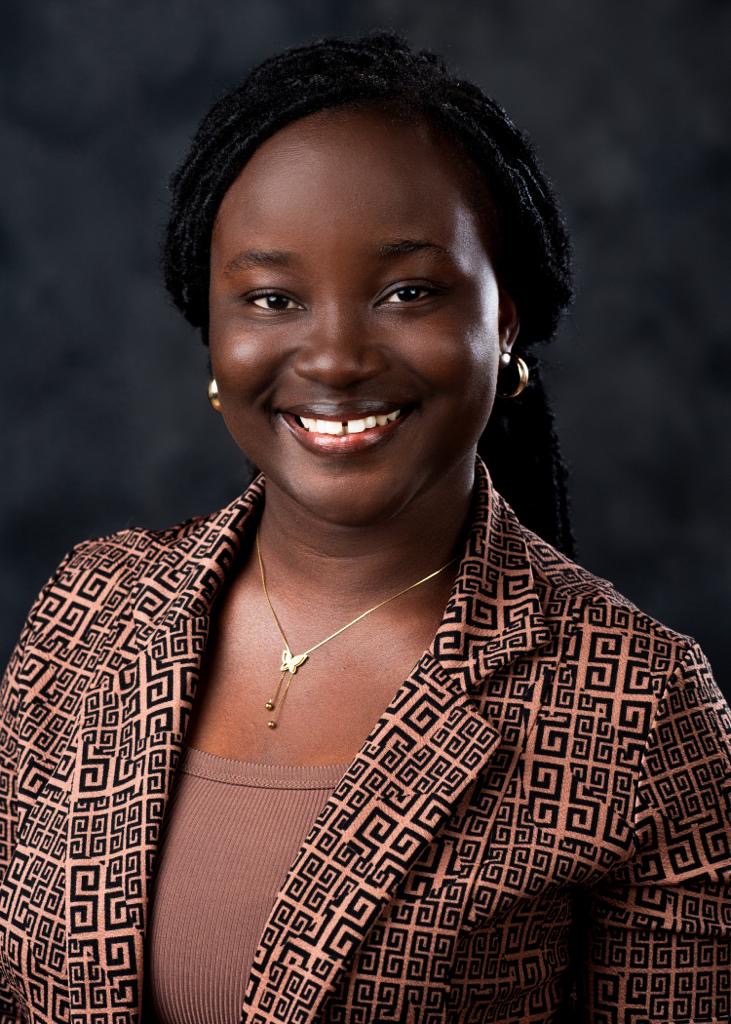 Headshot of Gladys Kainyah, wearing a brown blazer and standing in front of a gray backdrop