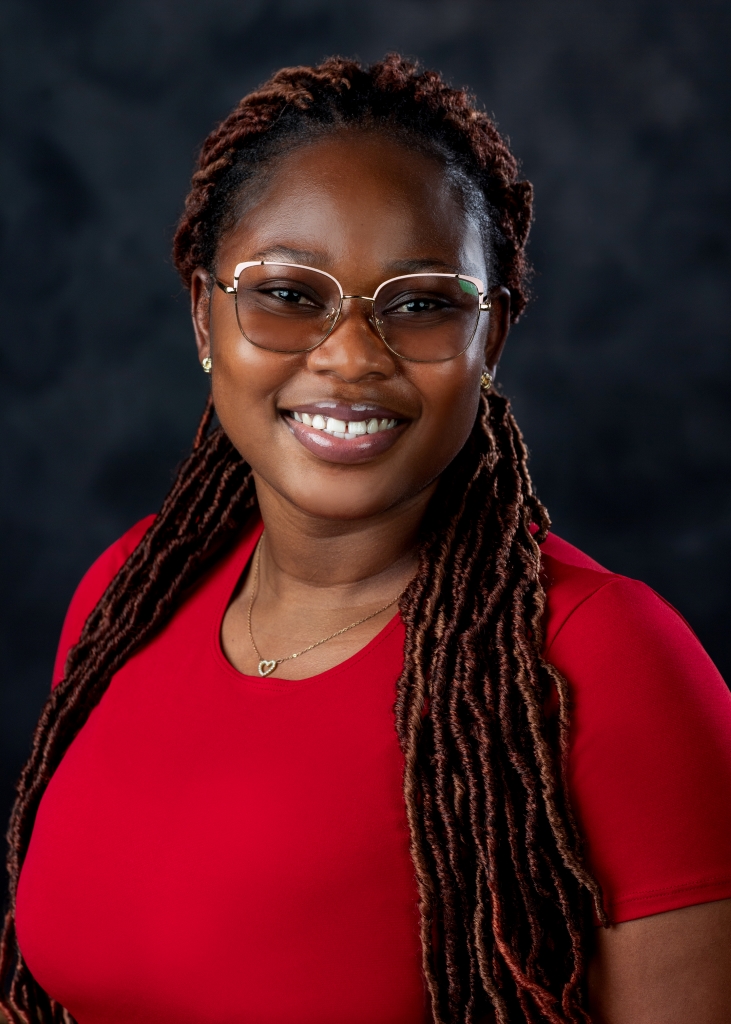 Photo graph of Justina Eshun, facing forward, wearing glasses and a red shirt, with a black background