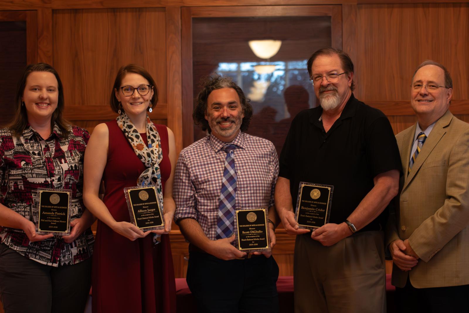 Five people, two women and three men, standing together; the four to the left hold award plaques.
