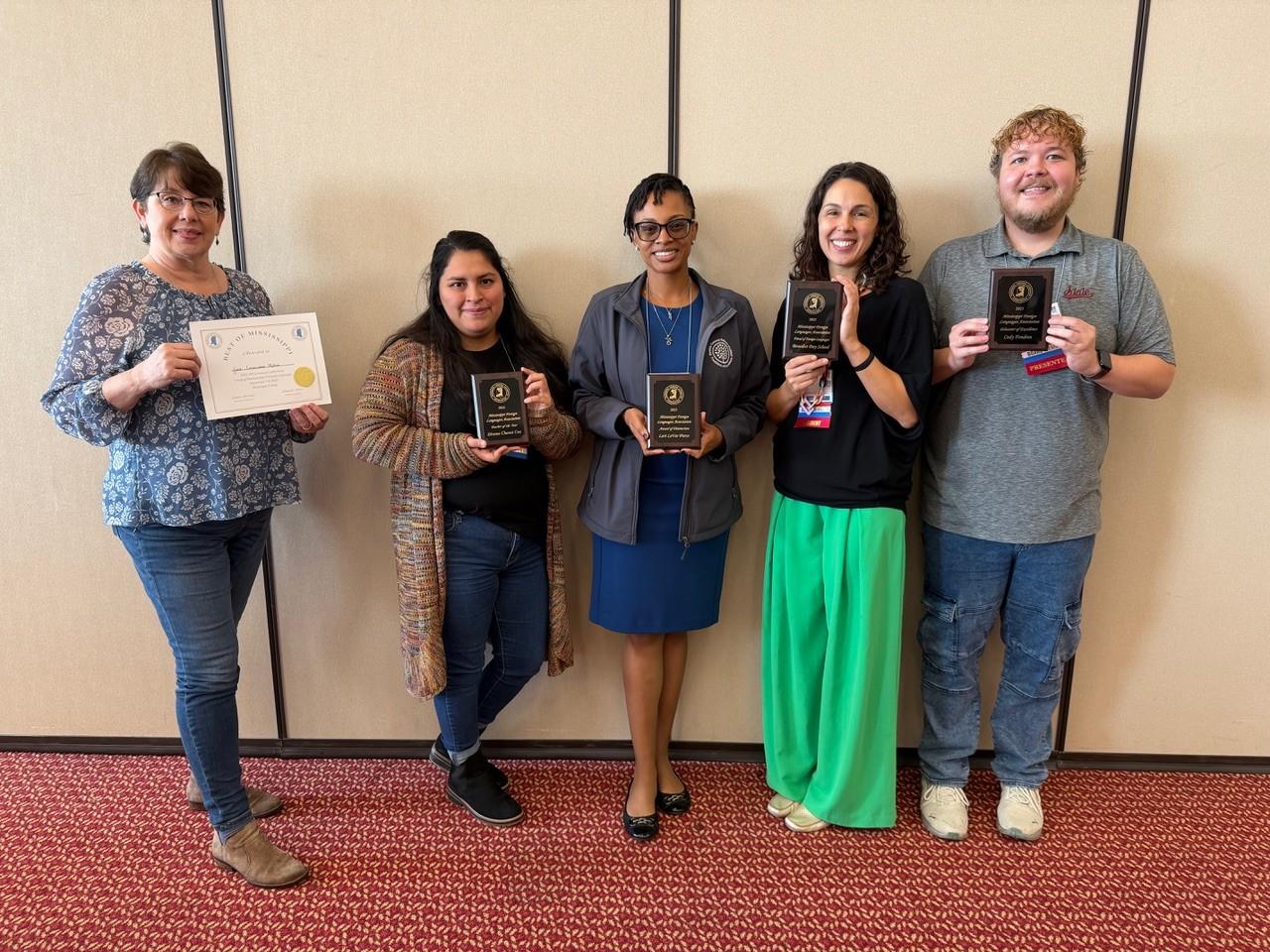 Five people standing in front of a beige wall holding awards