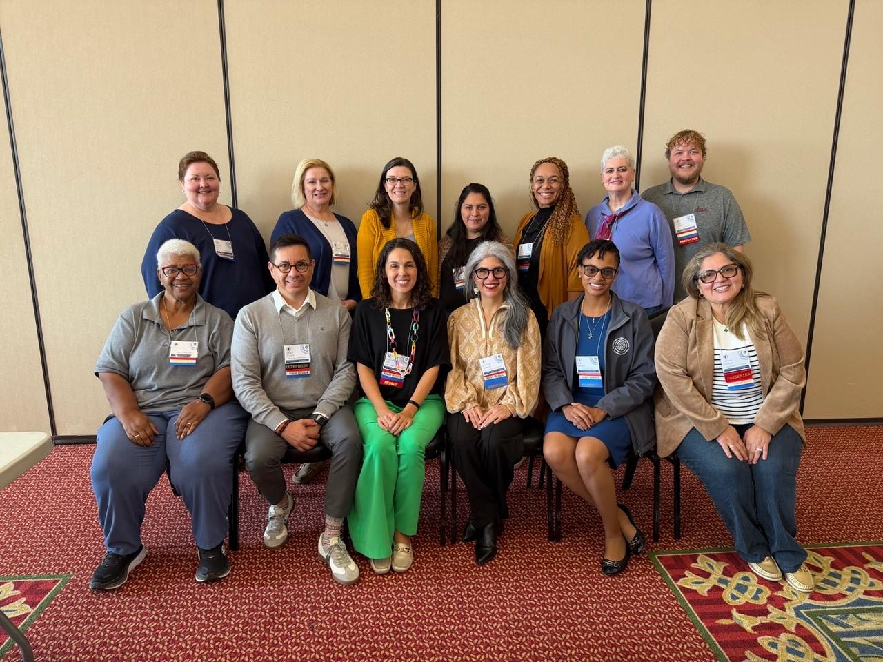 Seven people standing in front of a beige wall and six people sitting in chairs in front of them. Everyone is wearing conference badges with a red, white, and blue board member banner on the badge.