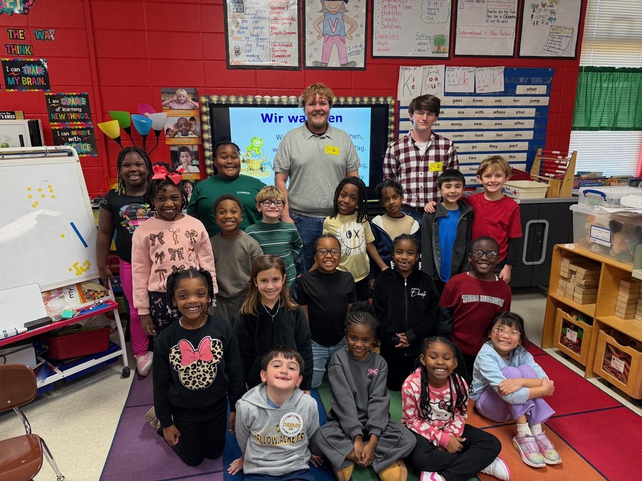 A group of first grade students standing on a carpet with two adults behind them.