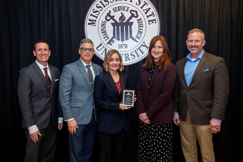 Five people, three men and two women, standing in front of the Mississippi State University seal; the woman in the middle holds an award plaque.