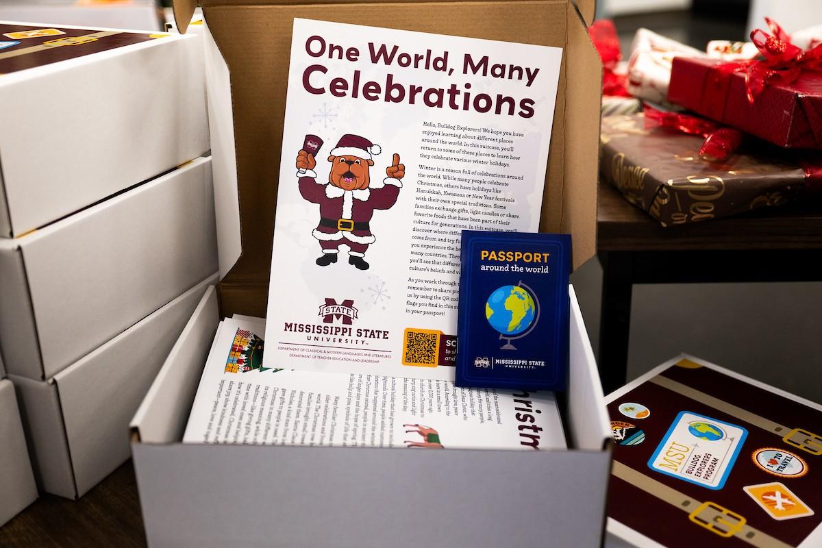 An open Bulldog Explorer Box with a sheet of paper inside that says “One World, Many Celebrations” with a Bully mascot wearing a maroon Santa outfit. There is also a card in the box that says “Passport around the world”.